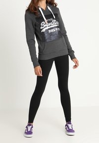 Felpa con cappuccio grigia con un logo in paillettes lucide, tasca frontale e cordini bianchi. Abbinata a leggings neri e scarpe da ginnastica viola.