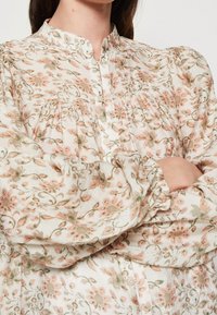 Kvinde iført en langærmet bluse med høj hals, med et pink og grønt blomster mønster, knapper langs fronten og puffede ærmer. Armene er korslagte.