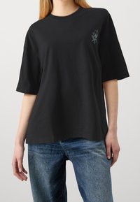 Svart bomullströja med lös passform, korta ärmar, rund halsringning och en liten vit broderad blommDetalj på bröstet.