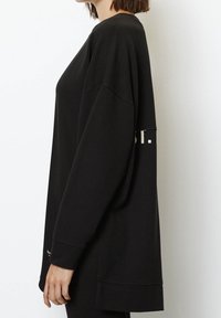 Mujer vestida con una camiseta negra de manga larga y oversize, con hombros caídos y un pequeño logo blanco cerca de la muñeca, mostrada de lado.