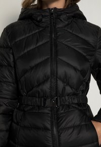 Una donna indossa un giubbotto imbottito nero con cappuccio, zip frontale e cintura elastica con una fibbia nera lucida.