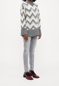 Graues und cremefarbenes Sweatshirt im Zickzackmuster mit hohem Kragen; kombiniert mit hellgrauen Jeans und glänzenden roten Mary Jane-Schuhen mit Blockabsatz.