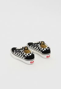 Paire de baskets Vans à motif damier noir et blanc avec lacets blancs et logo rouge sur semelle en caoutchouc blanche, vues de dos et de côté.