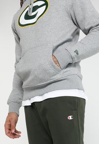 Sudadera gris con un logo verde y amarillo en el pecho, presenta un bolsillo delantero y puños acanalados, combinada con pantalones de chándal verde oscuro que muestran un logo.