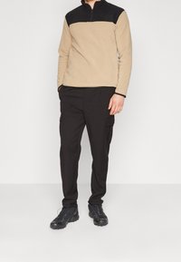 Sudadera de forro polar beige con panel negro en el pecho, cuello con cremallera, combinada con pantalones cargo negros con bolsillos laterales y zapatillas de deporte negras resistentes.