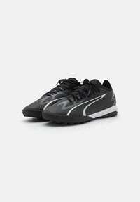 Par de zapatillas deportivas negras con diseño de zigzag y rayas blancas, cordones negros y suelas de goma texturizadas sobre un fondo blanco liso.