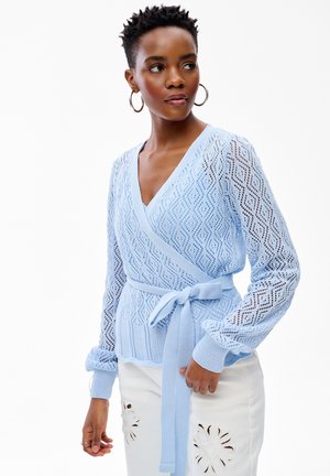 Kobieta w jasnoniebieskim, wzorzystym, dzianinowym swetrze z paskiem w talii i białych spodniach z wyciętymi kwiatowymi wzorami, patrząca w bok.