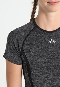 Lyhythihainen urheilupaita mustasta häilyvästä kankaasta, jossa on raglan-hihat ja pieni valkoinen logo rinnassa.