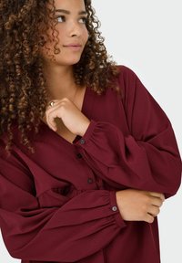 Burgundische Bluse mit V-Ausschnitt, Knopfverschluss und voluminösen Ärmeln. Strukturiert Stoff mit dezenten Falten an der Brust. Schwarze Knöpfe an den Ärmeln.