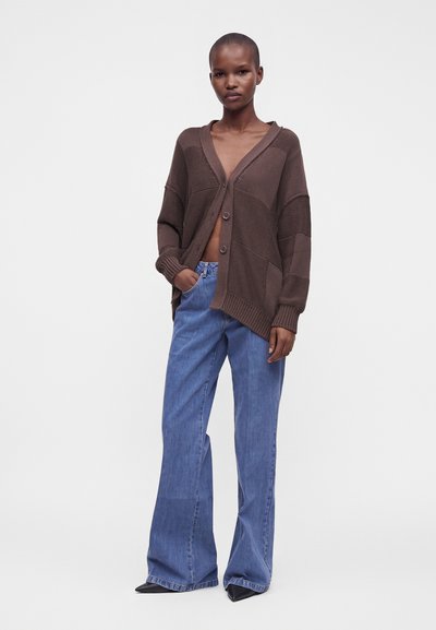Femme debout portant un cardigan marron ample boutonné et un jean bleu large avec des chaussures noires pointues sur un fond blanc uni.