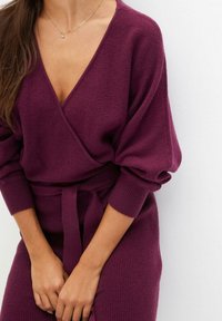 Pull-over croisé en bordeaux profond avec un décolleté en V, des manches larges et une ceinture assortie à la taille. Texture tricotée avec des poignets côtelés.