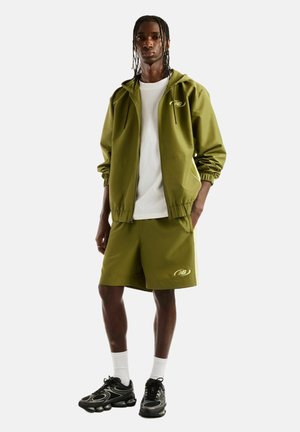 Jeune homme debout portant une veste à capuche New Balance vert olive et un short assorti avec un t-shirt blanc, des chaussettes blanches et des baskets noires.