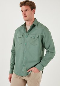 Cămașă button-up verde deschis, cu două buzunare la piept, butoane contrastante și guler clasic; realizată dintr-un material moale și texturat.