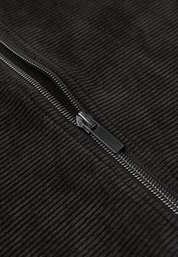 Zwarte corduroy stof met verticale ribbels, voorzien van een gladde metalen rits langs het midden. De ritspull is slank en rechthoekig.