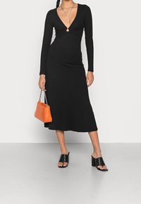 Musta ribbimekko, jossa on pitkät hihat, syvä V-pääntie ja avainaukko-detail vyötäröllä; yhdistetty oranssiin käsilaukkuun ja mustiin korkokenkiin.