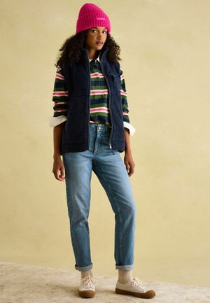 Navy fleece vest over a striped long-sleeve shirt, light blue jeans, cream sneakers, and a pink knit beanie. Soft textures and casual fit.