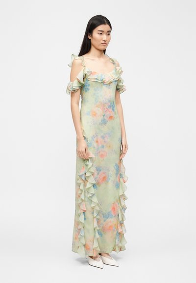 Γυναίκα που φοράει ένα μακρύ, ανοιχτοπράσινο φλοράλ φόρεμα με βολάν στα μανίκια που αφήνουν τους ώμους ακάλυπτους και βολάν κατά μήκος των πλευρών, συνδυασμένο με λευκά μυτερά παπούτσια.