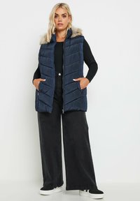 Meripaitainen puhviliivi, jossa on karvavuorattu huppu, vaakasuoralla tikkaustyylillä ja sivutaskuilla, yhdistettynä leveälahkeisiin mustiin farkkuihin ja lenkkareihin.