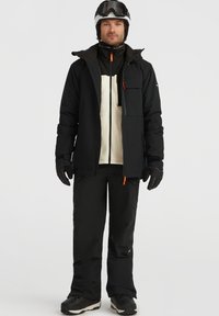 Zwarte skijack met een lichtgrijze fleece voering, voorzien van meerdere zakken en een oranje ritslip, gecombineerd met zwarte skibroeken en handschoenen.