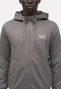 Homme portant un sweat à capuche gris zippé avec cordons et logo EA7 Emporio Armani blanc sur la poitrine gauche.