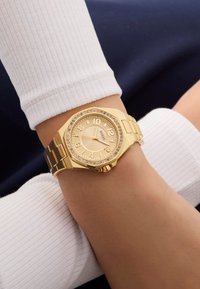 Zlaté náramkové hodinky s osmiúhelníkovým ciferníkem, textura zlatého ciferníku, arabské číslice a krystalové akcenty kolem lunety na zlatém kovovém pásku.
