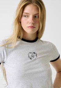 Szara koszulka z czarnym kołnierzykiem typu ringer. Na przodzie znajduje się czarne logo w kształcie serca. Wykonana z miękkiego, elastycznego materiału.