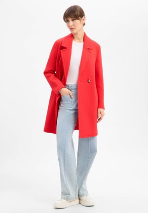 Frau steht und trägt einen leuchtend roten Mantel über einem weißen Hemd, hellblaue Jeans und weiße Sneakers vor einem schlichten weißen Hintergrund.