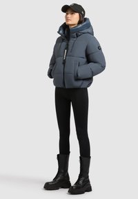 Donkerblauwe pufferjack met hoge kraag en ritssluiting. Voorzien van twee voorzakken, een verstelbare capuchon en een merklabel. Gemaakt in combinatie met zwarte leggings en veterschoenen.