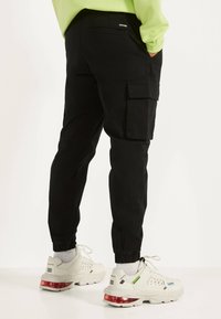 Mustat cargo-housut, joissa on joustavat hihan päät, sivutaskut ja slim fit -leikkaus, yhdistettyinä tukeviin valkoisiin tennareihin, joissa on punaisia yksityiskohtia ja teksturoitu pinta.