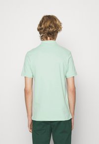 Mintgroene polo met korte mouwen, eenvoudig ontwerp en een klassieke kraag. G gedragen met donkergroene broek, gezien van achteren.