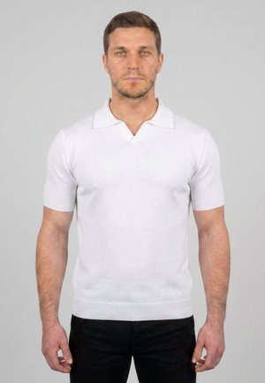 Hombre con cabello corto y castaño que lleva una camiseta polo blanca de manga corta y pantalones negros, de pie frente a un fondo liso de color gris claro.