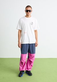 T-shirt blanche avec impression graphique sur la poitrine, associée à un pantalon baggy gris et rose. Baskets noires avec accents bleus.