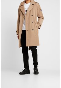 Béžový trenčkot s dvojitým zapínáním, velkým límcem a černými knoflíky. Nosený přes bílé tričko a černé kalhoty, doplněný černými botami.