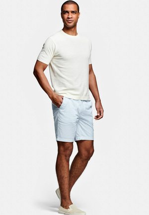 Mann steht mit einer Hand in der Tasche, trägt ein weißes kurzärmliges Strickshirt, hellblaue Shorts und beigen Slipper vor einem weißen Hintergrund.