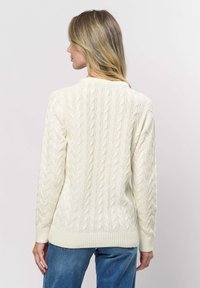 Woman with blonde hair wearing an off-white cable knit sweater and blue jeans, shown from the back against a plain background.