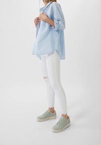 Camisa azul claro con botones y mangas remangadas, combinada con vaqueros pitillo blancos rotos y zapatillas verdes claras. Diseño simple y moderno.