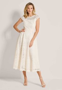 Weißes Spitzenkleid mit tailliertem Oberteil, kurzen Ärmeln und schwingendem Midirock, das mit floralen Mustern und einem gewellten Saum versehen ist.