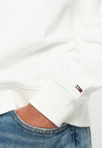 Osoba v bílém dlouhém rukávu s malým červeným, bílým a modrým logem na manžetě, ruka částečně v přední kapse modrých džín.