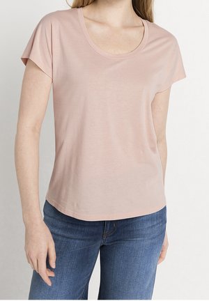 Mujer con una camiseta lisa de manga corta de color rosa claro y jeans azules, de pie frente a un fondo blanco.