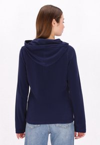 Felpa con cappuccio in pile blu navy, dal texture morbido, con cappuccio profondo e maniche lunghe, abbinata a jeans azzurri chiari. Nessun motivo visibile o dettagli in metallo.
