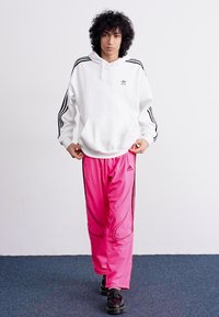 Hoodie branco com riscas pretas nas mangas, combinado com calças de fato de treino rosa brilhante com detalhes pretos. O modelo usa ténis pretos.