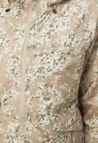 Tarnfarbende Jacke in Beige mit glatter Textur, ausgestattet mit einem Reißverschluss vorne und einer verstellbaren Kapuze. Grünes und beiges gesprenkeltes Muster überall.