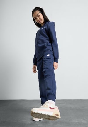 Dunkelblaue Sweatjacke mit Reißverschluss und passende Jogginghose aus weichem Stoff, mit einem weißen Nike-Logo. Sneakers haben pinke und beige Akzente.