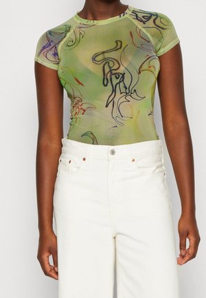 Personne portant un haut vert transparent à manches courtes avec des motifs de lignes abstraites et un pantalon blanc taille haute sur un fond uni.