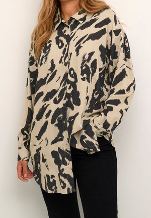 Kvinde iført en beige skjorte med knapper med sorte abstrakte penselstrøg og sorte bukser, den ene hånd i lommen.