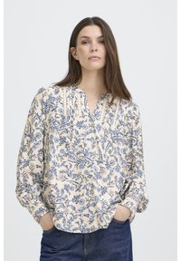 Blouse à motifs floraux en crème avec des dessins bleus. Dotée d'un col en V, de plis et de manches longues avec des poignets froncés. Texture douce.