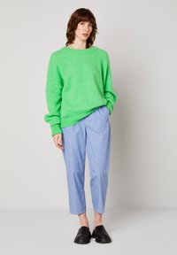 Noor naine kannab erkrohelist liialdatud suuruses kampsunit, siniste ja valgete triipudega pükse ning musti libisevaid jalatseid, seisab halli seina taustal.