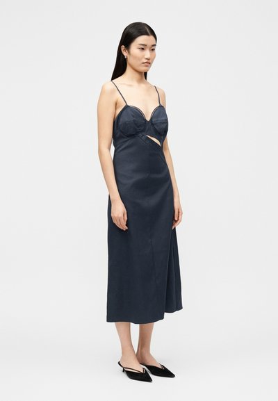 Γυναίκα που φοράει σκούρο μπλε μίντι φόρεμα με λεπτές τιράντες, ενσωματωμένα στηθόδεσα, πλαϊνή κοψίματα και μαύρα μυτερά παπούτσια slingback.