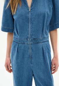 Denim-Overall mit kurzen Ärmeln, Reißverschlussverschluss, taillierter Passform, Falten an der Taille und Kontraststeppnähten in einem mittelblauen Farbton.