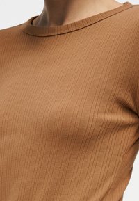 Κοντινό πλάνο ενός ατόμου που φοράει καφέ ριγωτό μακρυμάνικο τοπ με στρογγυλή λαιμόκοψη, δείχνοντας την υφή του υφάσματος και τη εφαρμογή στο άνω μέρος του κορμού.